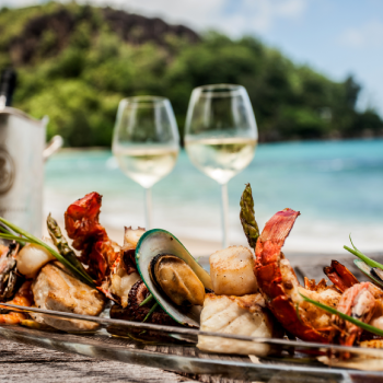 Eksklusiv seafood‑anretning med hummer, muslinger og asparges serveret ved stranden på Seychellerne – to glas hvidvin og turkisblåt hav i baggrunden skaber en luksuriøs, tropisk spiseoplevelse.